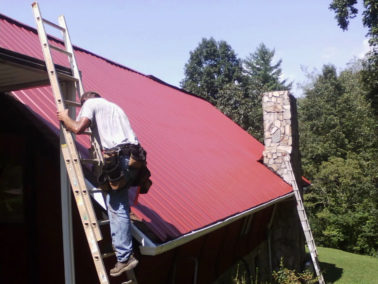 Roofer on a ladder beside a red metal roof and stone chimney — Soffit & Fascia Repair by Roofing Brothers in Pflugerville